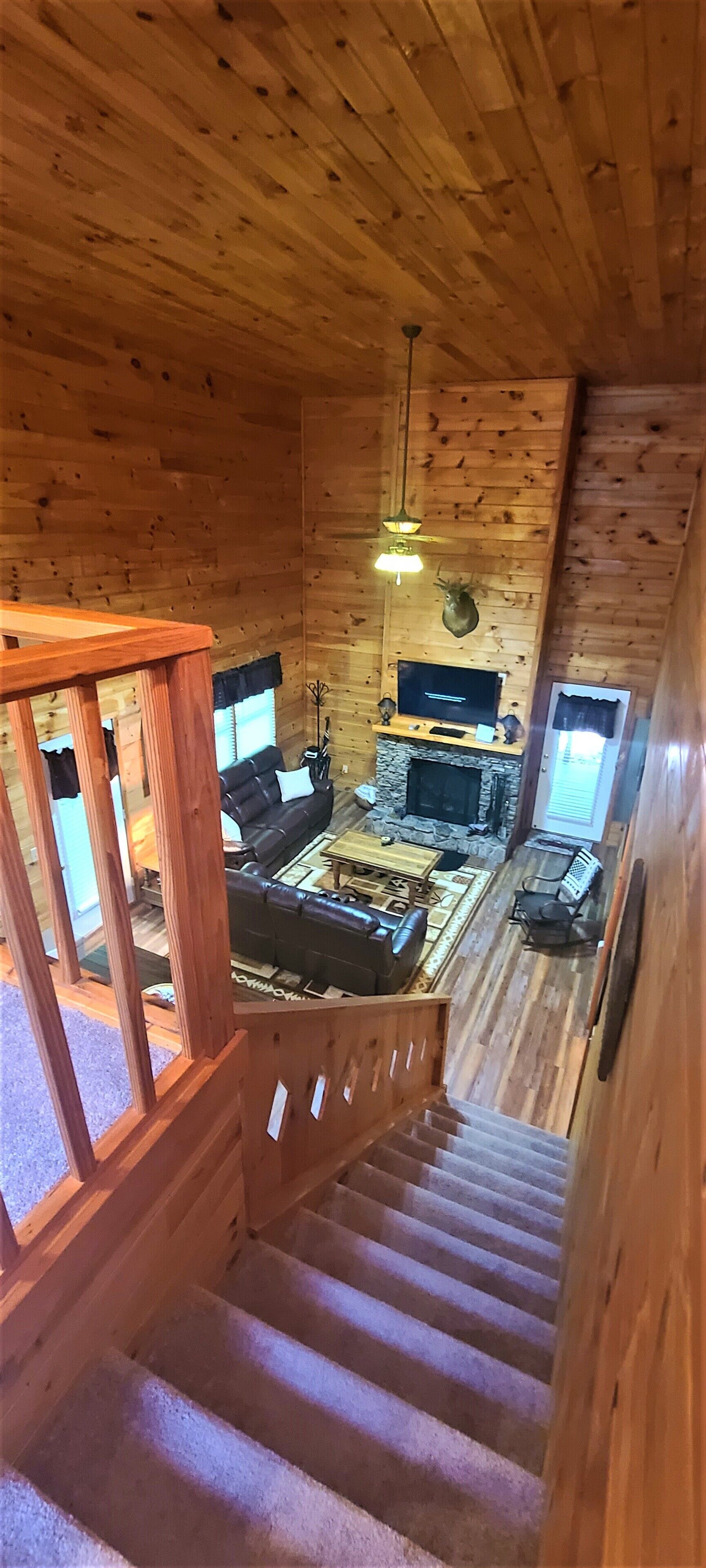 A wooden cabin interior with a stone fireplace, a black leather sofa, and a wooden staircase.