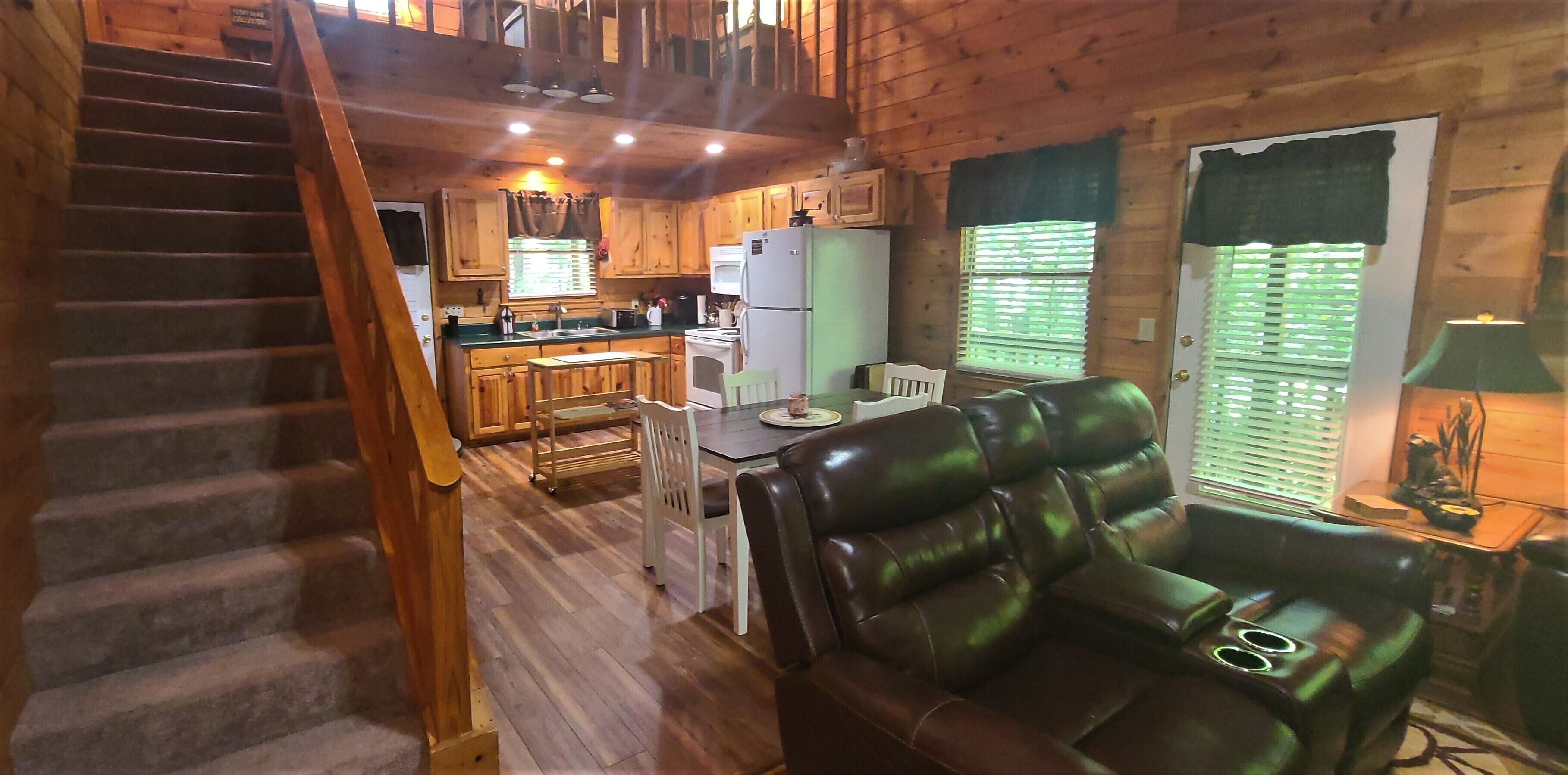 Interior de una cabaña con escalera, pisos de madera, zona de comedor con mesa y sillas, sofá de cuero, cocina con muebles y electrodomésticos de madera, y ventana con persianas.