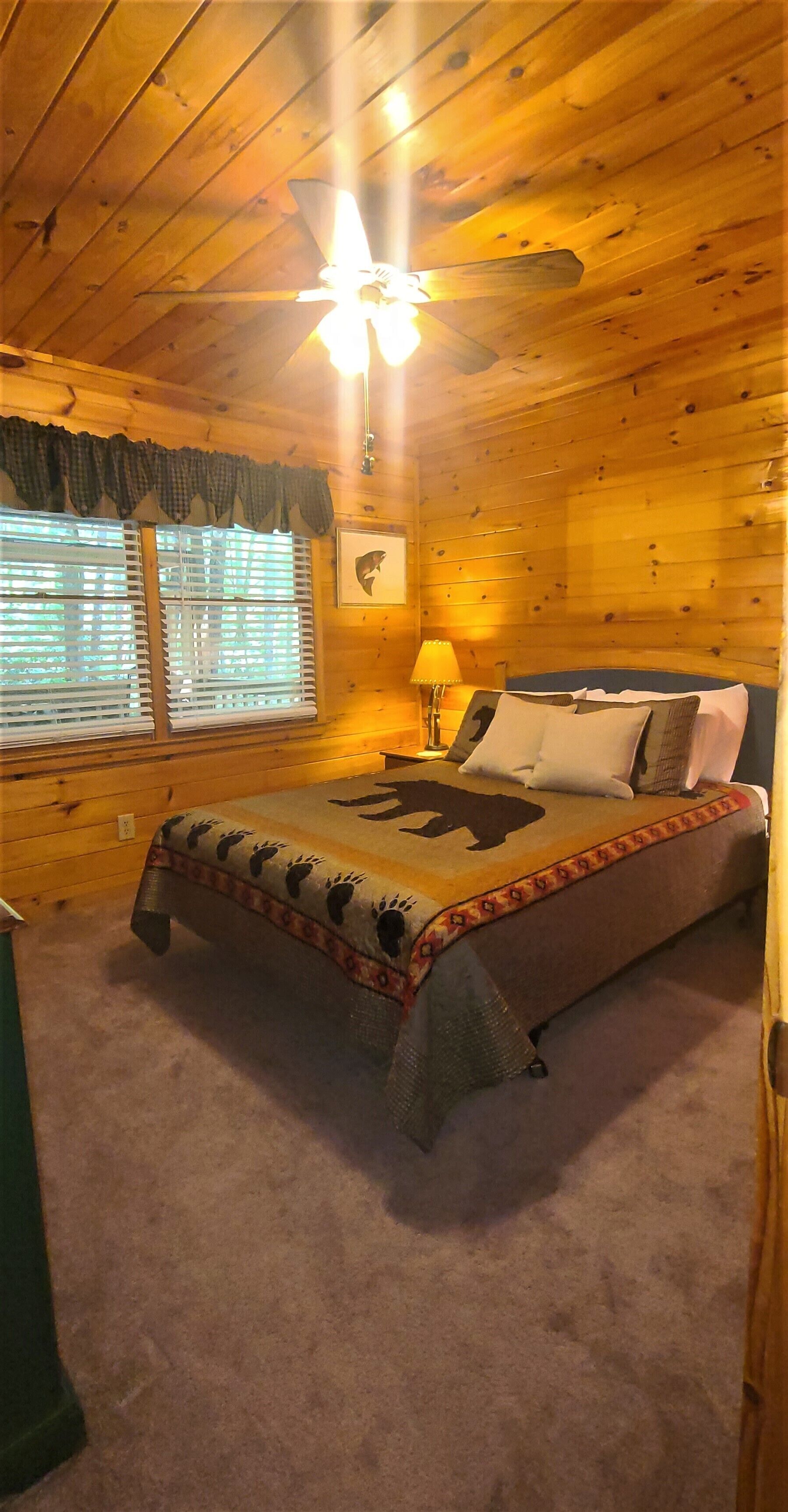 Ein Schlafzimmer einer Holzhütte mit Bett, Deckenventilator, Fenster mit Jalousien und Nachttisch mit Lampe.
