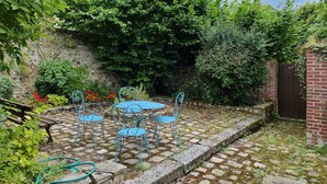 A garden patio with a table and chairs, surrounded by lush greenery and a stone wall.