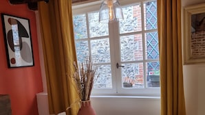 A room with a round white table, a chair, a vase with dried plants, a window with curtains, and a framed picture on the wall.