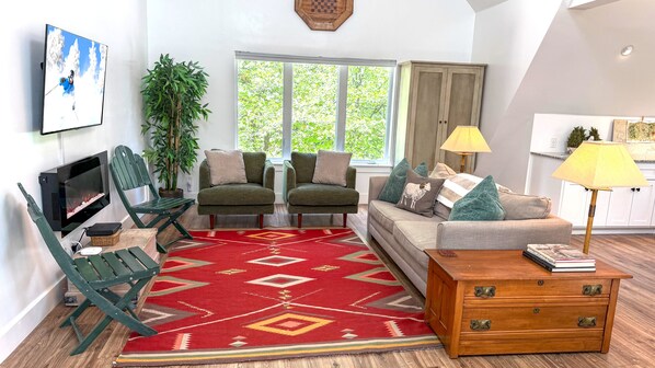 Una sala de estar con televisor, dos sillones, un sofá, un aparador de madera, un tapete rojo y un ventanal con vista a árboles.