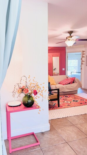 A living room with a pink-framed desk, a vase with flowers, a ceiling fan, a sofa with cushions, and a patterned rug.