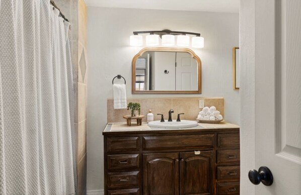 A bathroom with a wooden vanity, a large mirror with lights, a towel rack, and a door with a doorknob.