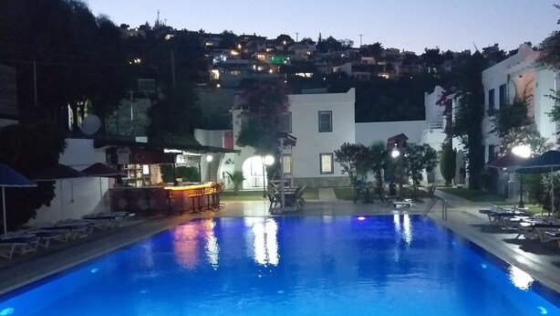 Una piscina con un borde de baldosas y sillas de descanso, rodeada de edificios y árboles.