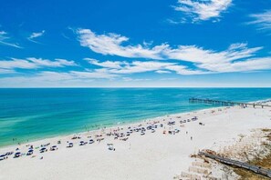 Une plage avec une jetée qui s’avance dans l’eau, un ciel dégagé et de nombreux parasols sur le sable.