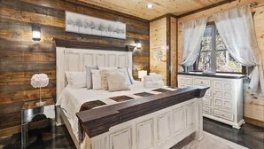 A bedroom with a wooden ceiling, a bed with white bedding, a nightstand, a dresser, and a window with curtains. - Three Wildflowers Cabin - cozy retreat for up to 6 (Broken Bow)