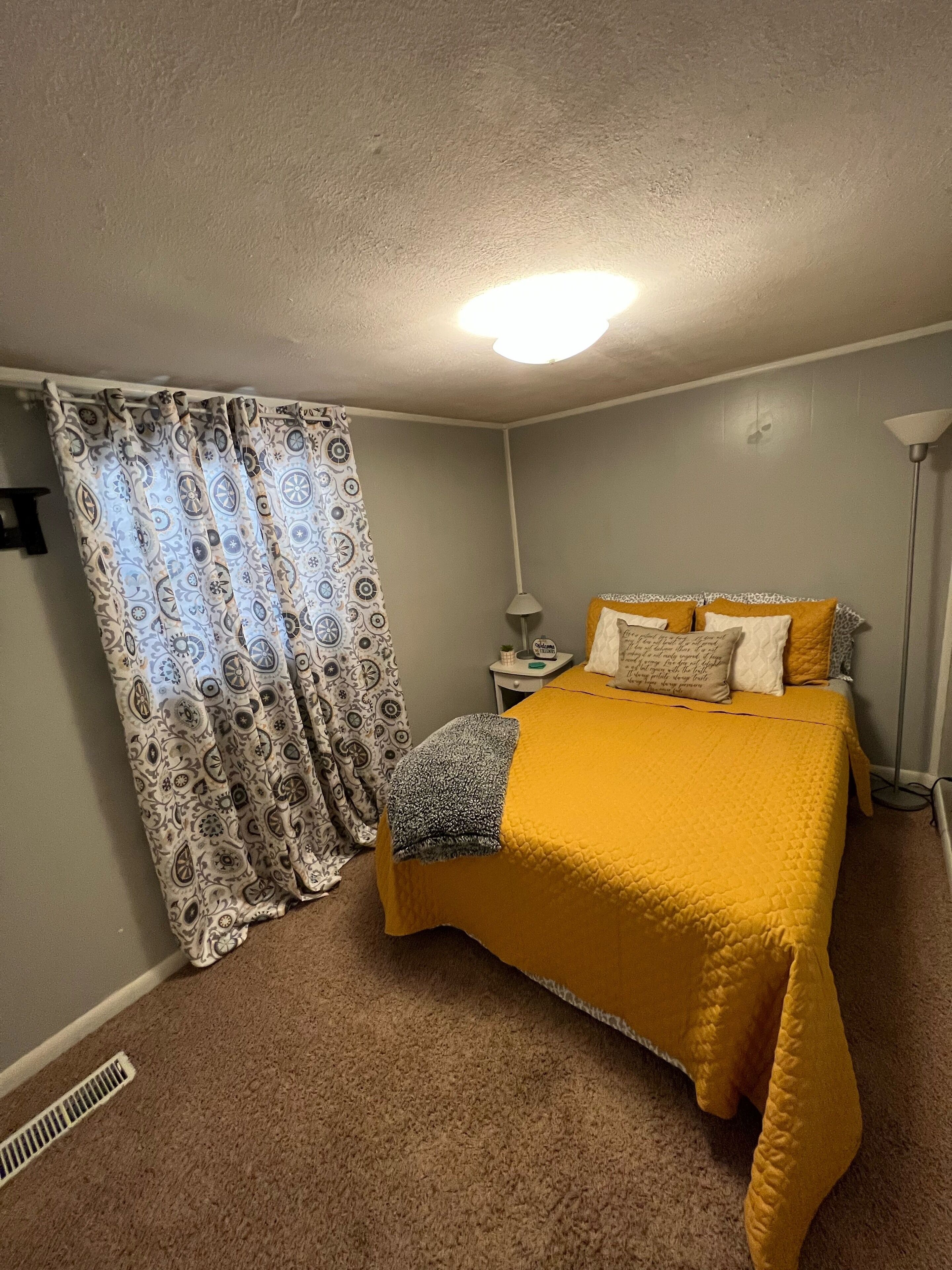 Quarto com cama, mesa de cabeceira, luminária e cortina estampada.