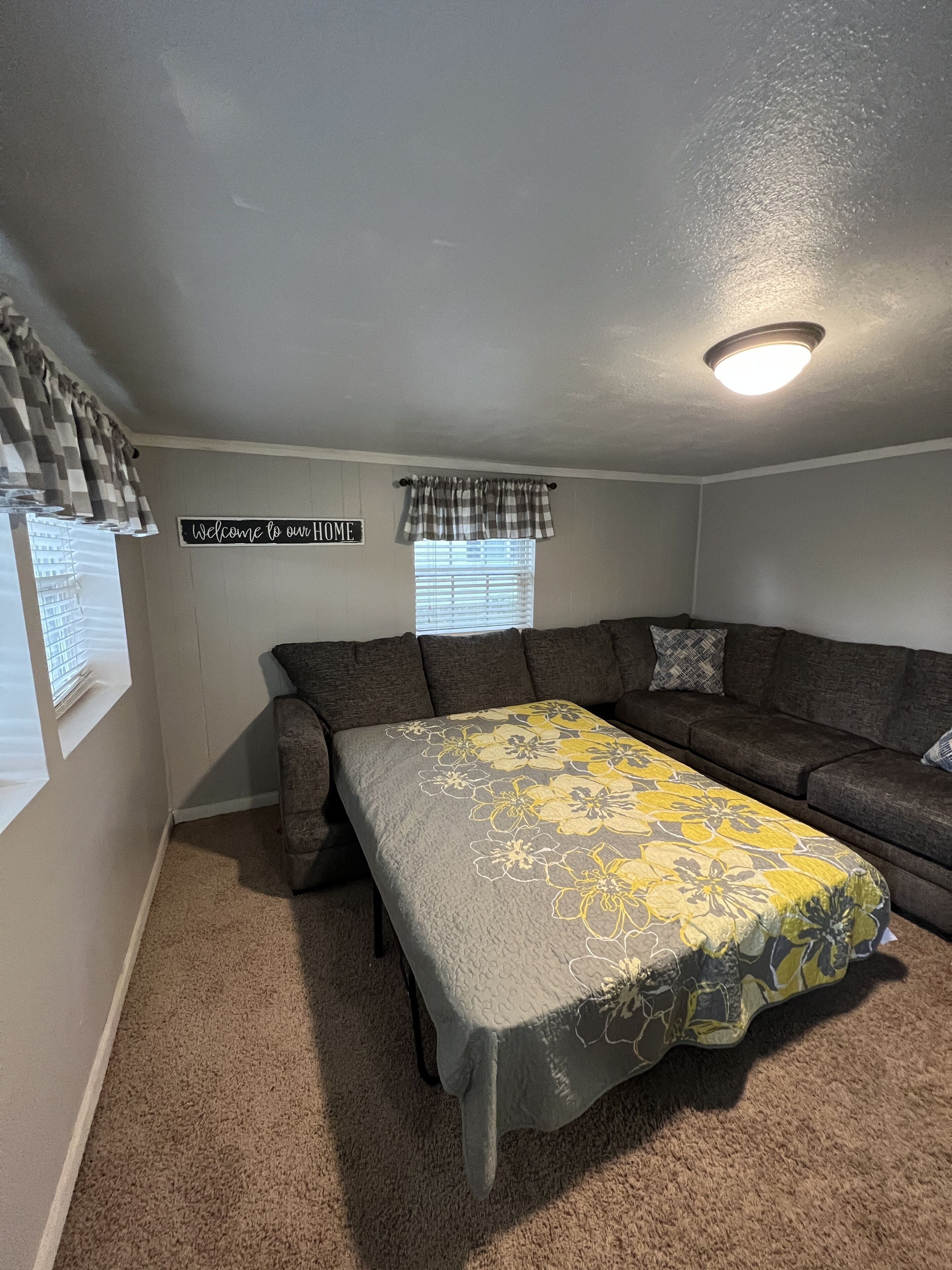 A cozy living room with a sectional sofa, a large bed with a floral bedspread, a 'Welcome to our home' sign, and a window with blinds.