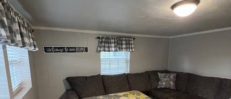 A cozy living room with a sectional sofa, a large bed with a floral bedspread, a 'Welcome to our home' sign, and a window with blinds.