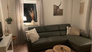 A modern living room with a dark green sofa, two round wooden tables, and a window with sheer curtains. - Vacation house-Braunlage apartment 2 (Braunlage)