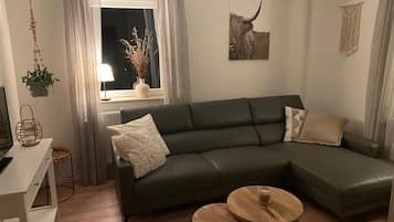 Ein modernes Wohnzimmer mit einem dunkelgrünen Sofa, zwei runden Holztischen und einem Fenster mit durchsichtigen Vorhängen.