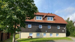 A two-story building with a dark exterior and a red-tiled roof. There are multiple windows on the upper floor and a small porch area with a bench. - Vacation house-Braunlage apartment 2 (Braunlage)