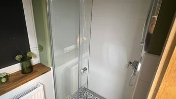 Une douche avec une porte en verre, un radiateur blanc, des carreaux de sol à motifs et une petite fenêtre agrémentée d’un vase.