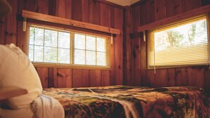 Una habitación de cabaña de madera con dos ventanas, una cama con colcha estampada y almohada blanca.