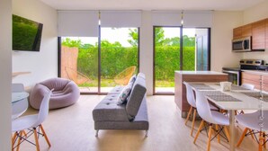 A modern living room with a grey sofa, white chairs, a glass dining table, and a kitchen area with wooden cabinets and a microwave. - Captivating 2-bed Apartment (Jacó)