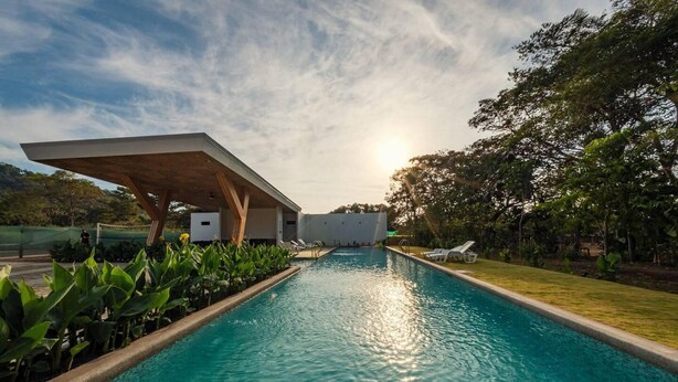 A swimming pool with a modern pavilion structure, surrounded by lush greenery and a clear sky. - Captivating 2-bed Apartment (Jacó)
