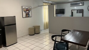 A compact kitchen with a refrigerator, a dining table with chairs, a small trash bin, and a wall-mounted air conditioner.