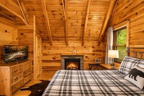 Ein Schlafzimmer in einer Blockhütte mit Kamin, einem Bett mit kariertem Überwurf, einem Fernseher auf einer Kommode und einem Fenster mit Jalousien.