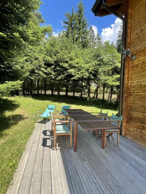 A wooden deck with a table and chairs, surrounded by a lush green garden and tall trees.