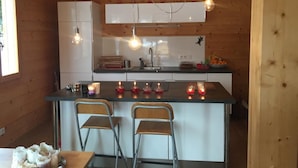 Een kleine keuken met houten wanden, een centraal keukenblok met barkrukken, hanglampen en een koelkast.