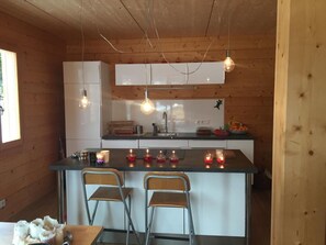 A compact kitchen with wooden walls, a central island with bar stools, pendant lighting, and a refrigerator.