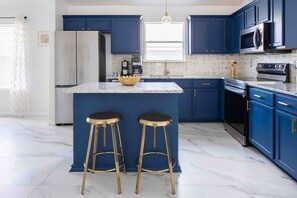 A kitchen with blue cabinets, a marble island, two gold stools, stainless steel appliances, and a window. - 🌵 Stylish Hot Tub Oasis 🌵 (San Antonio)