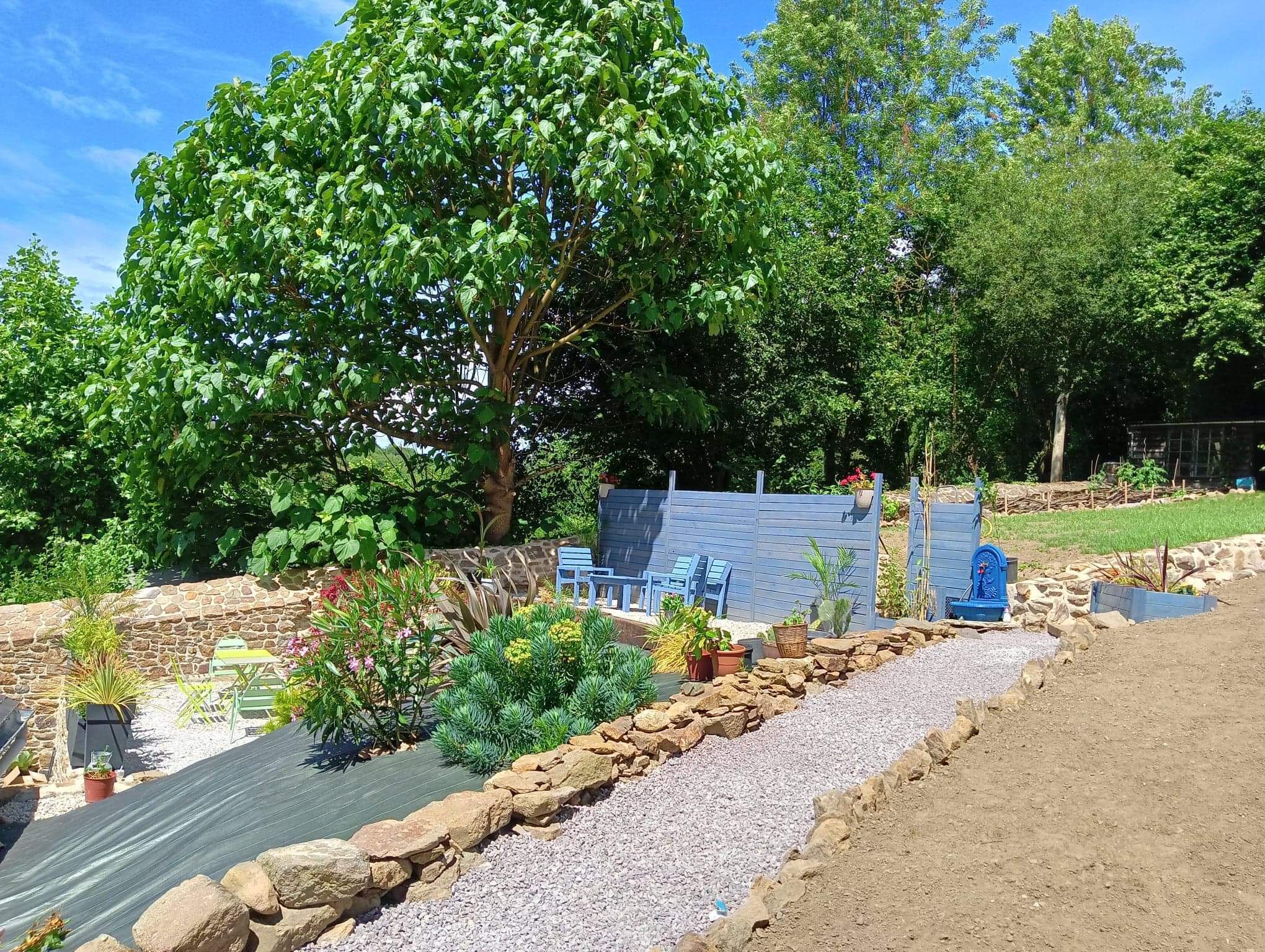 Um jardim com muro de pedra, banco de madeira e cerca azul.