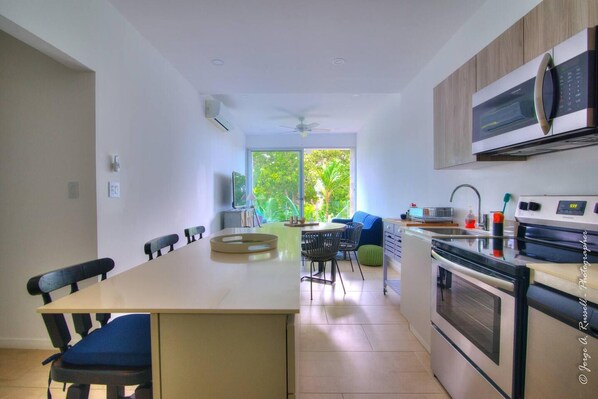 A modern kitchen with a central island, dining area, and a view of greenery outside. - Beautiful Small Three Bedroom (Jacó)