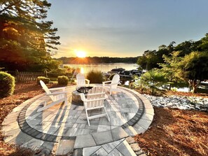 A circular outdoor seating area with Adirondack chairs and a fire pit, overlooking a lake with boats docked and a sunset in the background. - Modern Lakefront Oasis - Private Pool & Salt Water HotTub with Beautiful Sunsets (Charlotte)