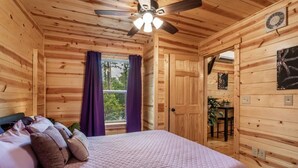 Habitación de cabaña de madera con cama, cortinas moradas, ventilador de techo y ventana con vista a árboles.
