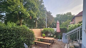 Un patio con mesa y bancos de madera, rodeado de follaje denso y una escalera de metal.