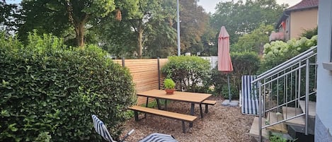 A patio area with a wooden table and benches, surrounded by dense foliage and a metal staircase.