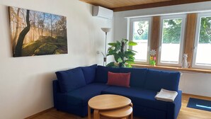 Un salón con un sofá azul, una pequeña mesa de madera y un ventanal con vistas a árboles.