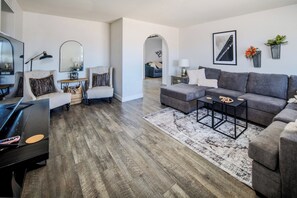 A modern living room with a grey sofa, armchairs, a coffee table, and a piano. There are two lamps, a rug, and a framed picture on the wall. - Come with the Crew: Game Room, Bunks & Style! (Lake Havasu City)