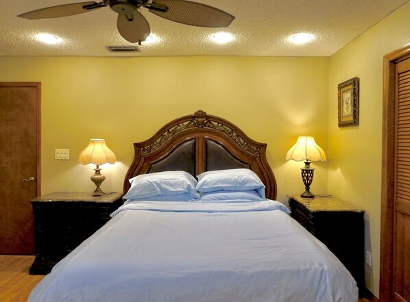 A neatly made bed with a wooden headboard, two bedside lamps, and a ceiling fan in a room with yellow walls and a wooden door.