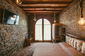 A rustic room with a stone wall, a bed with a white bedspread, a wooden bench, and a TV mounted on the wall.