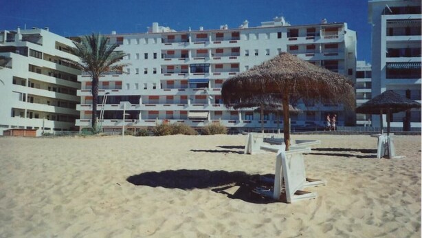 Una playa con un paraguas de paja y un hotel de varios pisos al fondo.