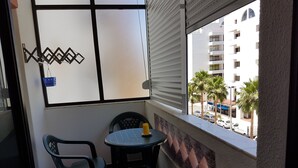 Un pequeño balcón con una mesa y dos sillas, una ventana con persianas y vistas a un paisaje urbano con edificios y palmeras.