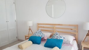 Una habitación de hotel con una cama de madera, dos mesitas de noche, un espejo redondo y un armario blanco.