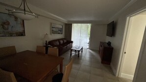 A living room with a brown sofa, a wooden coffee table, and a dining table with chairs. There is a painting on the wall, a lamp, and a television mounted on the wall. - Five minutes from disney world! perfect for family sleeps up to 5-6 with balcony (Orlando)