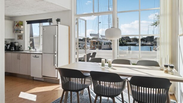 A modern kitchen with white cabinets, a refrigerator, and a dining area with a table and chairs. A large window offers a view of a marina with boats. - Stunning home in Ebeltoft with sauna (Ebeltoft)