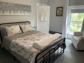 A neatly made bed with floral bedding, a bedside table, a chair, and a window with a view of greenery. - Historic Skye House in the Heart of Northport (Northport)