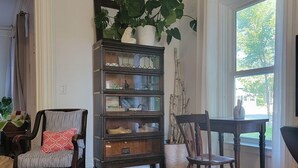Una habitación con piso de madera, un tapete estampado, una librería de madera oscura con puertas de vidrio, dos sillas y un gran ventanal con cortinas.