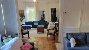 Un salón espacioso con suelo de madera, un sofá azul, un sofá gris y una mesa de centro de madera. Se ve una puerta que da a otra habitación al fondo.