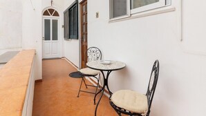 Un couloir étroit doté d’un sol carrelé, d’un mur blanc, d’une fenêtre avec des stores et d’une petite table ronde accompagnée de deux chaises.