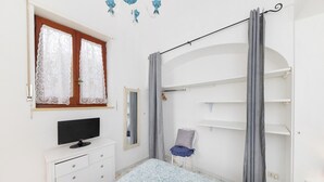 Une chambre à coucher avec un lit, une commode, un téléviseur, une chaise et une fenêtre avec des rideaux.