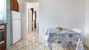 Une cuisine avec un sol carrelé, un réfrigérateur blanc, un meuble en bois et une table recouverte d’une nappe à motifs, sur laquelle se trouve un saladier rempli de citrons.
