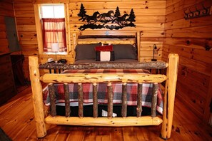 A rustic wooden cabin interior with a bed, a window with curtains, and a wall decoration of horses and trees.