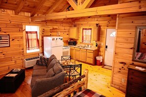Interior de una cabaña con sofá, mesa de comedor, zona de cocina y ventana con cortinas.